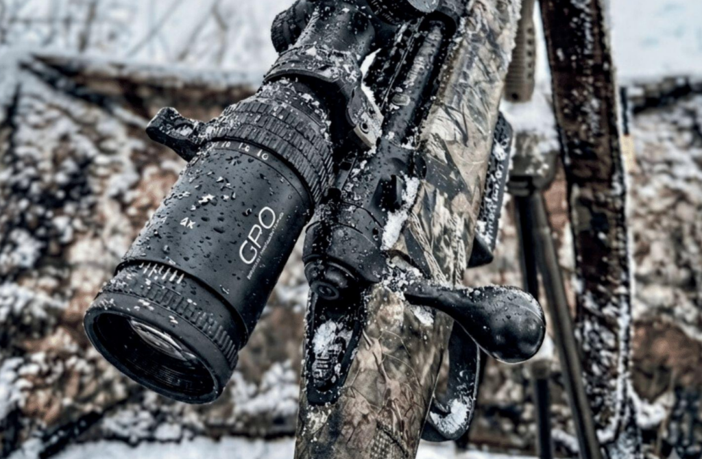 Close-up of a camouflage rifle with a black GPO scope, both covered in snow and water droplets, against a blurred snowy background.