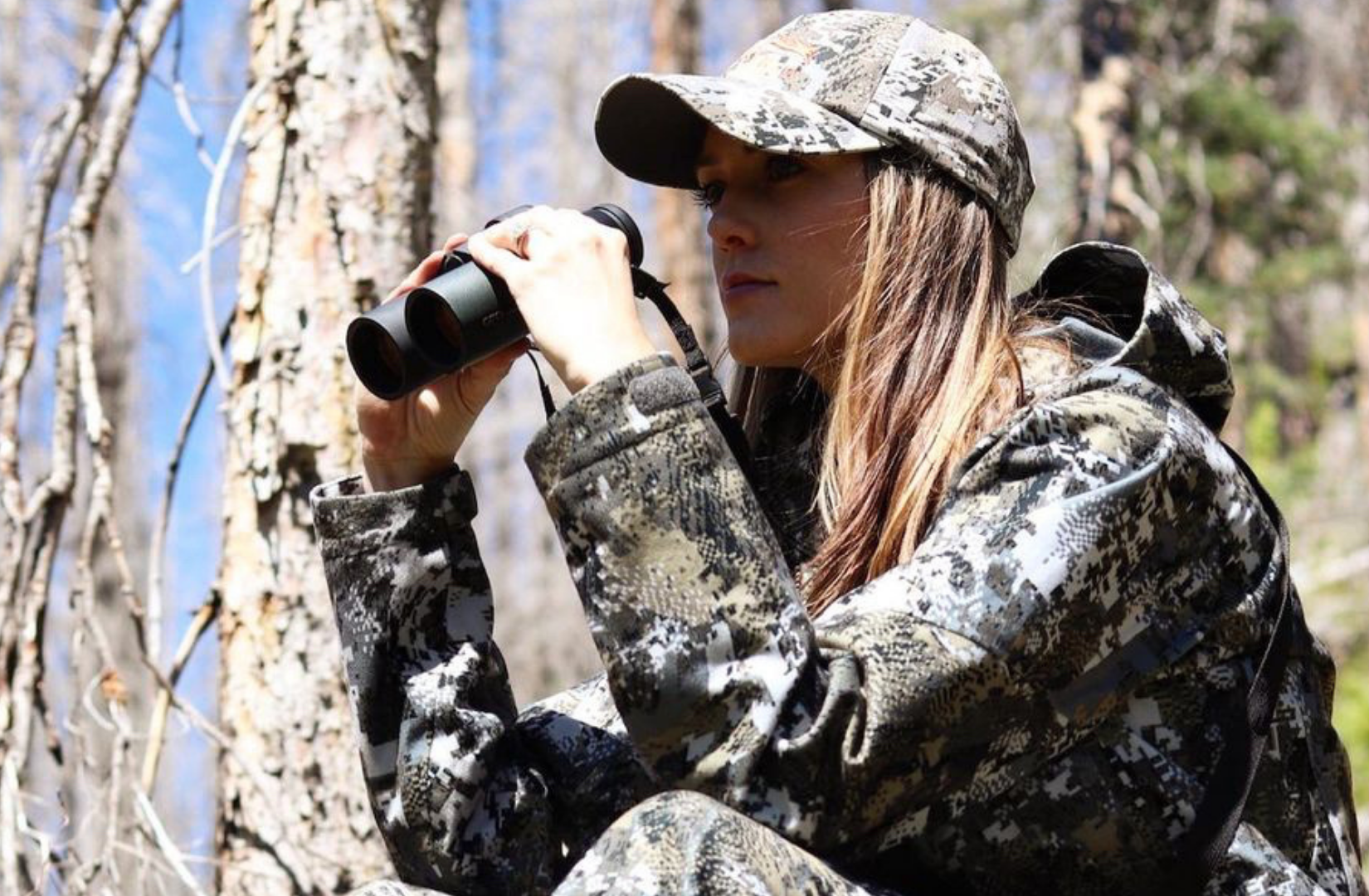A woman in camouflage clothing and a baseball cap looks through binoculars in a forest setting.
