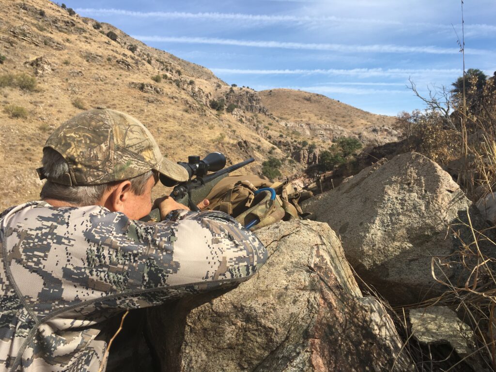 Man in camo looking over rocks through his long range scope on rifle.