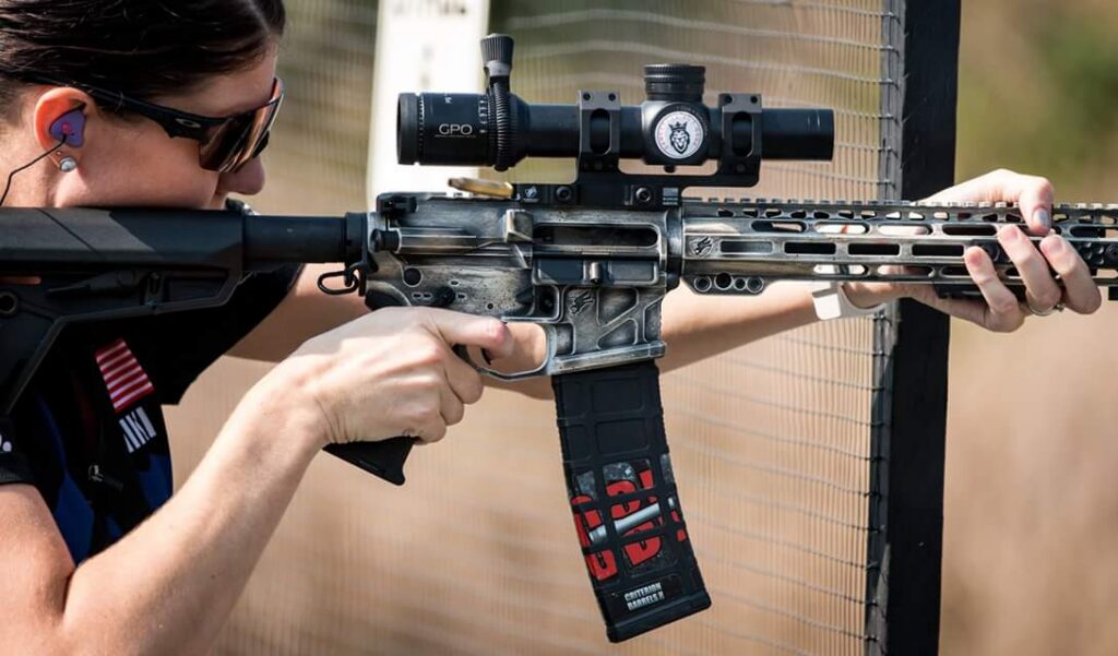 A woman wearing sunglasses and purple ear protection aims a distressed AR-style rifle with a scope, her hands gripping the weapon.