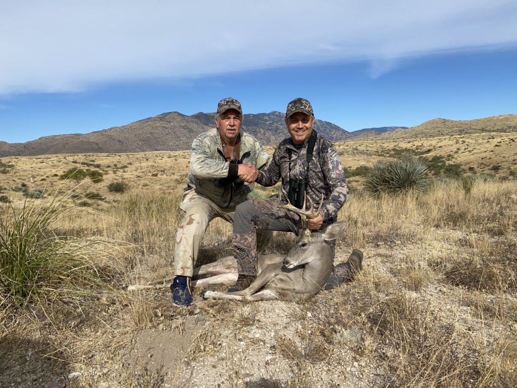 Two hunters in camouflage shake hands next to a harvested buck deer in a dry, mountainous outdoor setting.