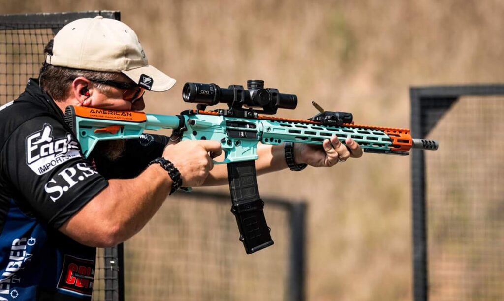 A man in a tan cap and sunglasses aims a teal and orange AR-style rifle with a scope at a shooting range, with a brass casing mid-air.