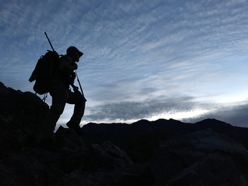 Silhouette of a person with a backpack and hat, hiking on rocky ground against a cloudy sky with mountains in the background.