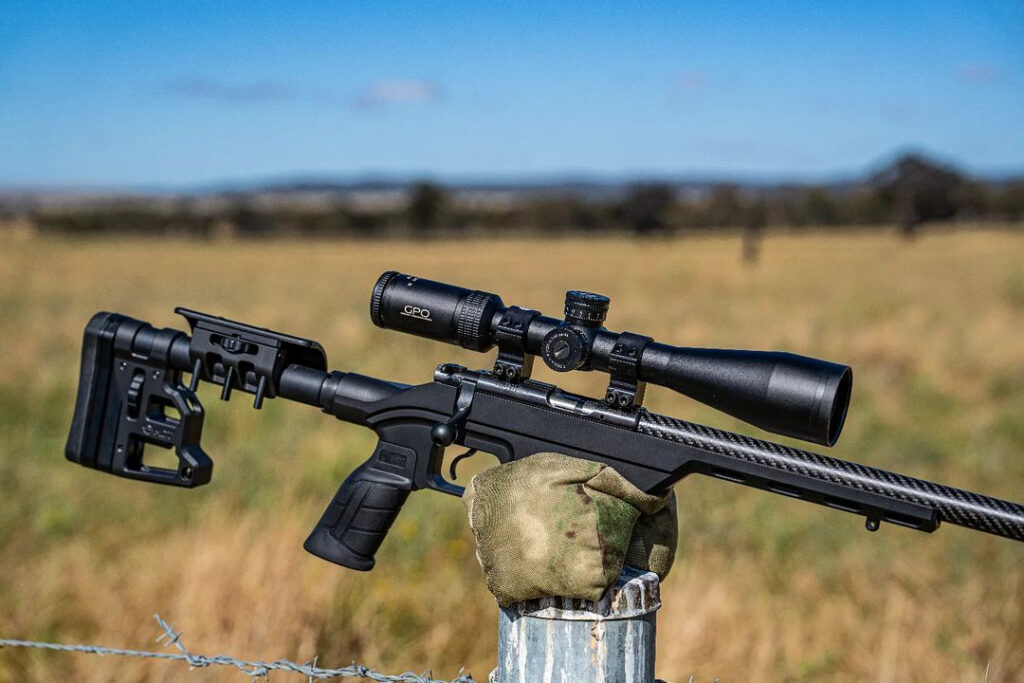 A black rifle with a scope rests on a camouflage sandbag on a metal fence post in a field under a blue sky.