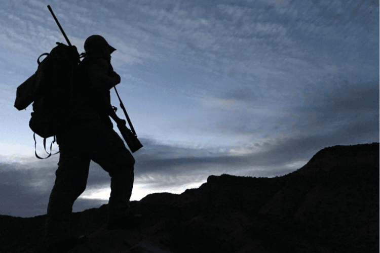 Silhouette of a person with a backpack and hat, hiking on rocky ground against a cloudy sky with mountains in the background.