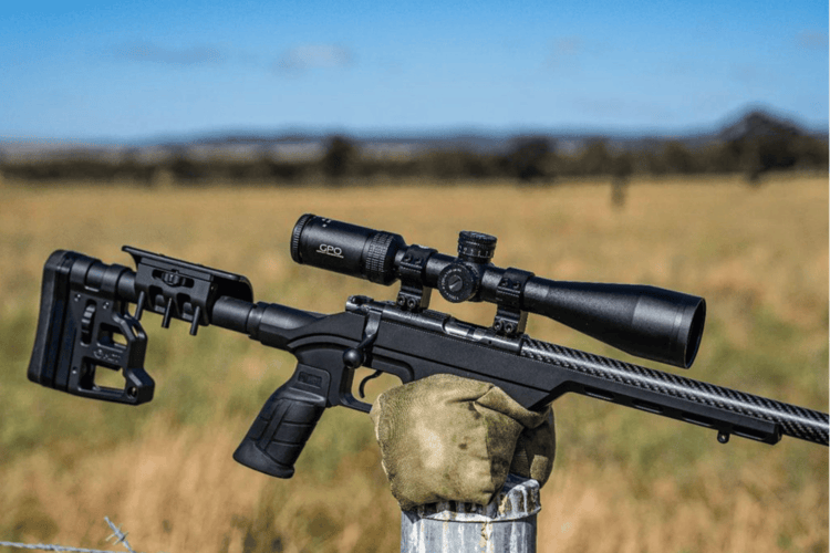 A black rifle with a scope rests on a camouflage sandbag on a metal fence post in a field under a blue sky.