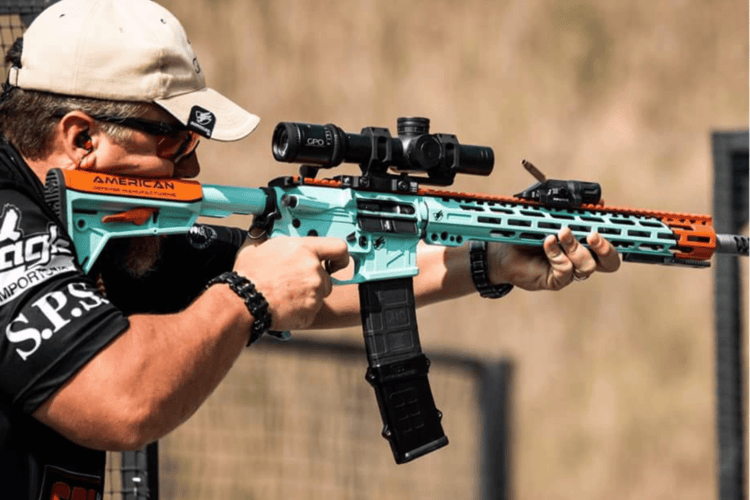 A man in a tan cap and sunglasses aims a teal and orange AR-style rifle with a scope at a shooting range, with a brass casing mid-air.