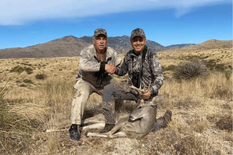 Two hunters in camouflage shake hands next to a harvested buck deer in a dry, mountainous outdoor setting.