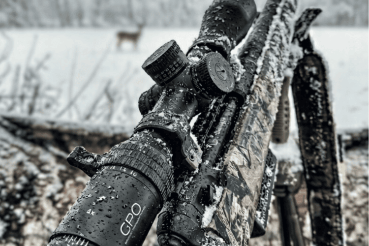 Close-up of a camouflage rifle with a black GPO scope, both covered in snow and water droplets, against a blurred snowy background.