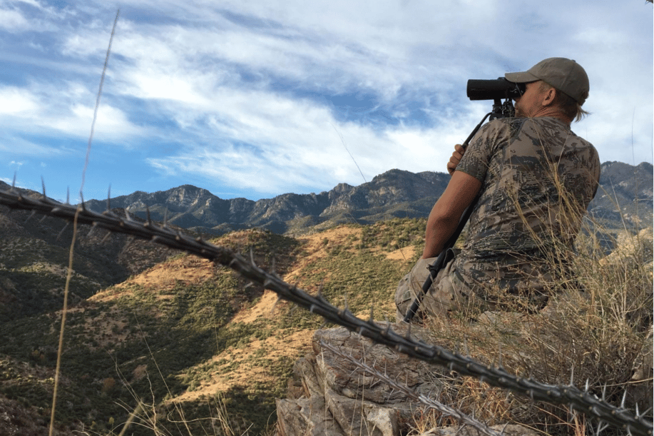 Man in camo sitting out in the feilds with a spotting scope looking for his next hunt